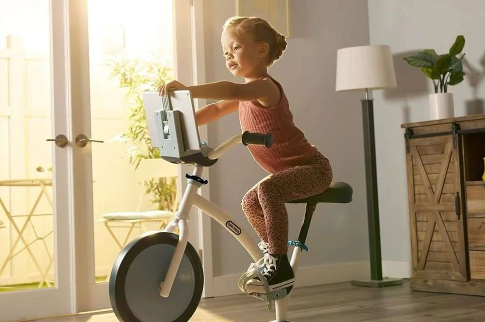 A young child is seated on a stationary exercise bike with a screen attachment, positioned indoors with natural light streaming in.