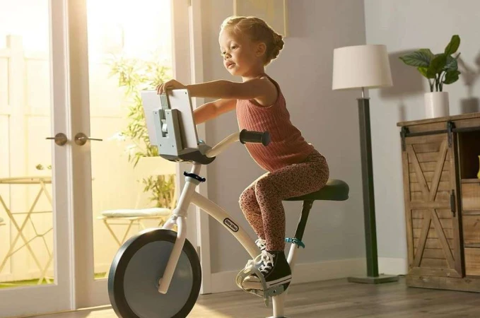 A young child is seated on a stationary exercise bike with a screen attachment, positioned indoors with natural light streaming in.