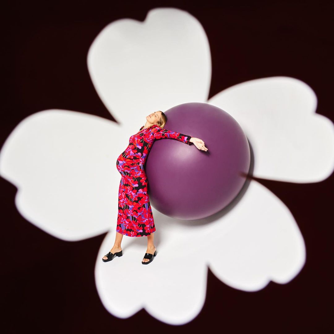 Pregnant woman in floral dress leaning against purple sphere in front of an abstract flower on a dark brown background. Maternity portrait.