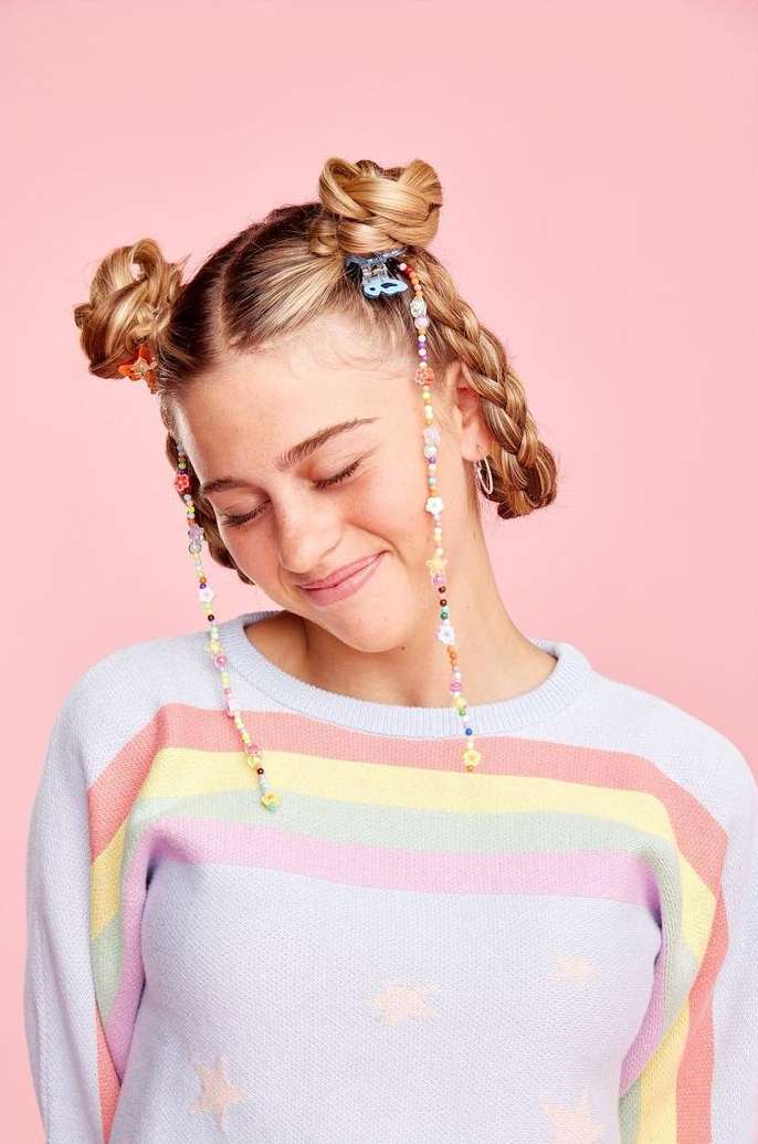 A young woman with space buns and beaded hair accessories smiles softly against a pink background, wearing a rainbow-striped sweater.
