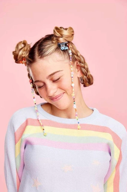 A young woman with space buns and beaded hair accessories smiles softly against a pink background, wearing a rainbow-striped sweater.