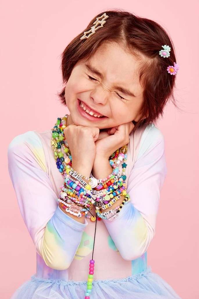 A young child with closed eyes, smiling, and wearing numerous colourful beaded bracelets and necklaces against a pink background in a close-up shot.
