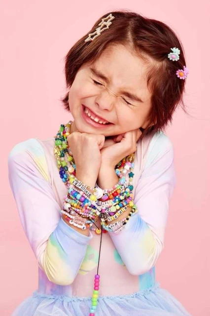A young child with closed eyes, smiling, and wearing numerous colourful beaded bracelets and necklaces against a pink background in a close-up shot.