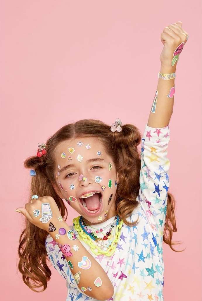 A happy child with stickers all over her face and arms raises her fist in a celebratory gesture against a solid pink background.