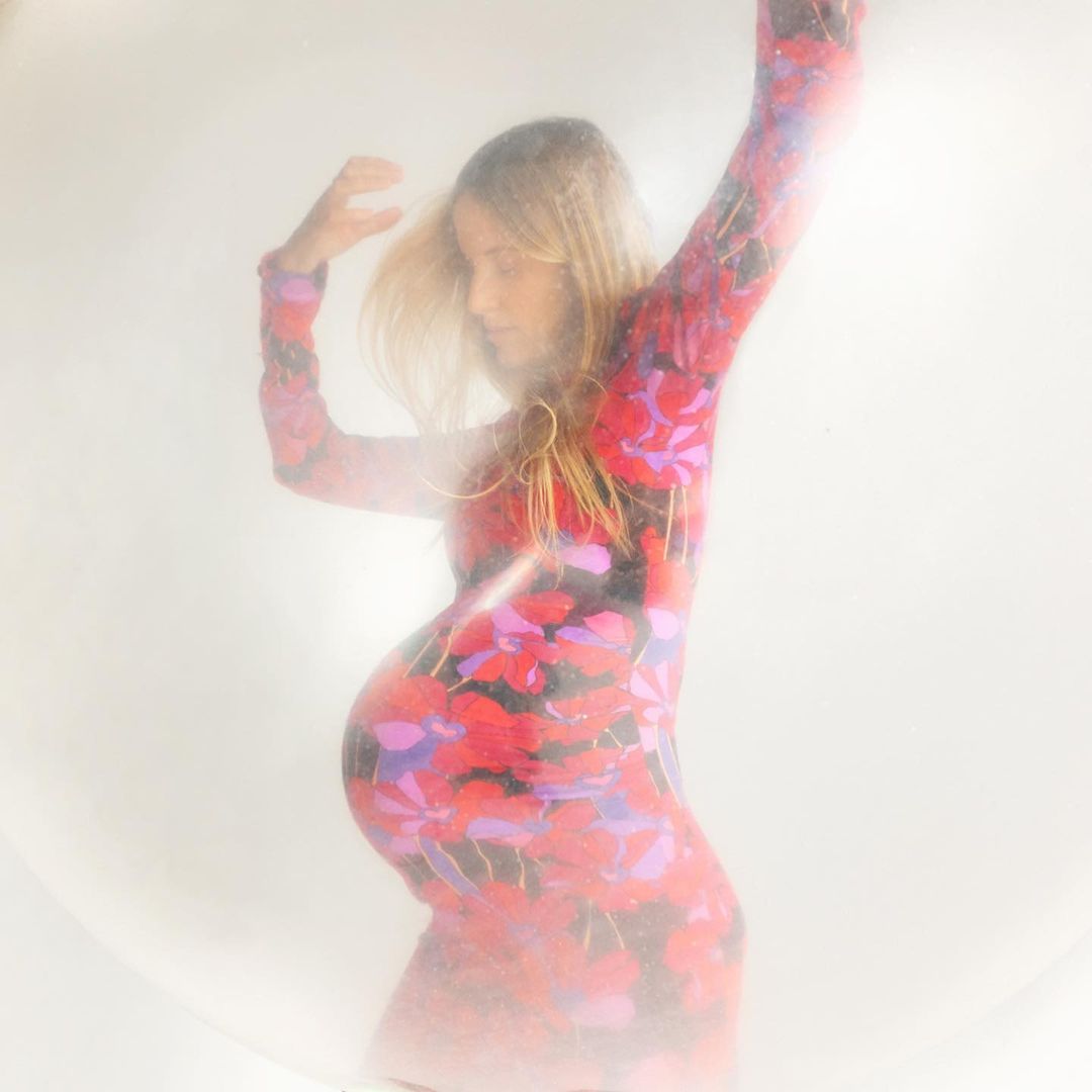 Pregnant woman poses in a floral-patterned dress, standing in front of a diffused, bright background, creating a soft and dreamy aesthetic.