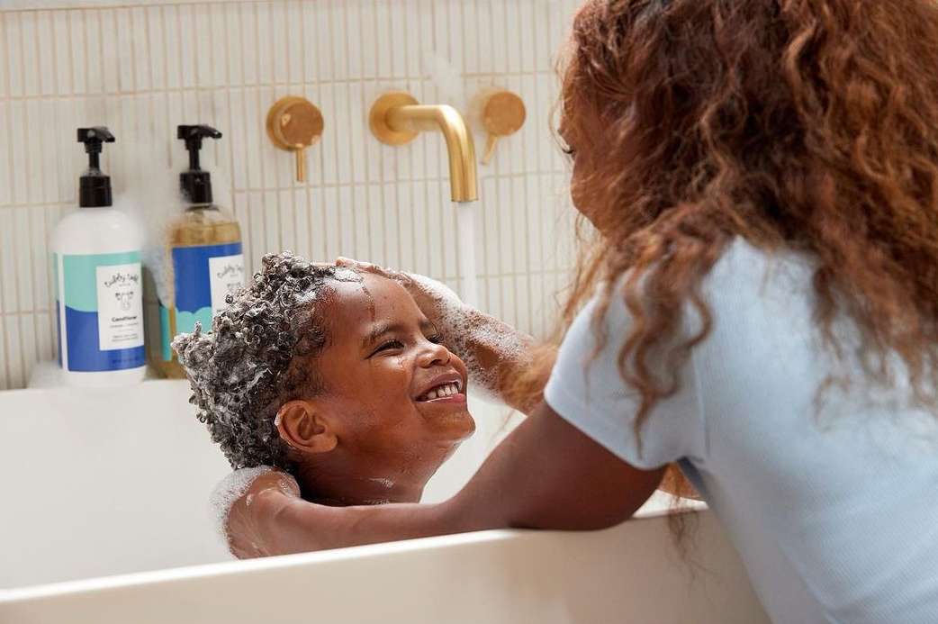 A happy child gets hair washed in a white tub by an adult with brown hair, featuring hair care product bottles and a gold faucet.
