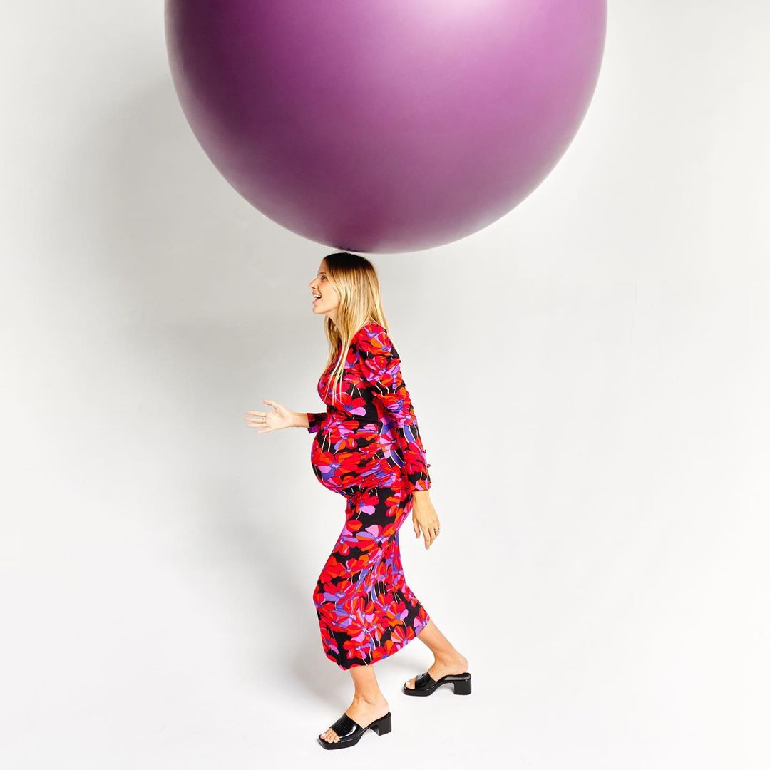 Pregnant woman in floral dress walking under a large purple sphere in a studio setting, promoting maternity fashion and related content.
