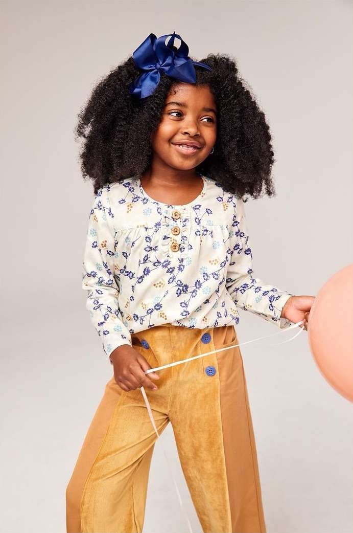 Young girl with dark curly hair smiles wearing a floral top, tan pants, and navy bow. She holds a peach balloon in a light studio.