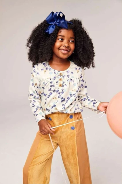 Young girl with dark curly hair smiles wearing a floral top, tan pants, and navy bow. She holds a peach balloon in a light studio.