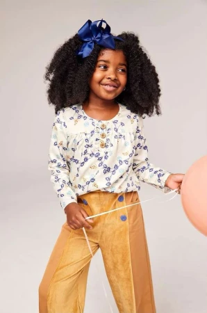 Young girl with dark curly hair smiles wearing a floral top, tan pants, and navy bow. She holds a peach balloon in a light studio.