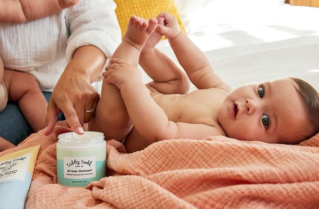 Infant lies on a blanket as an adult applies cream to their leg. Product packaging is visible. Soft, natural light fills the scene.