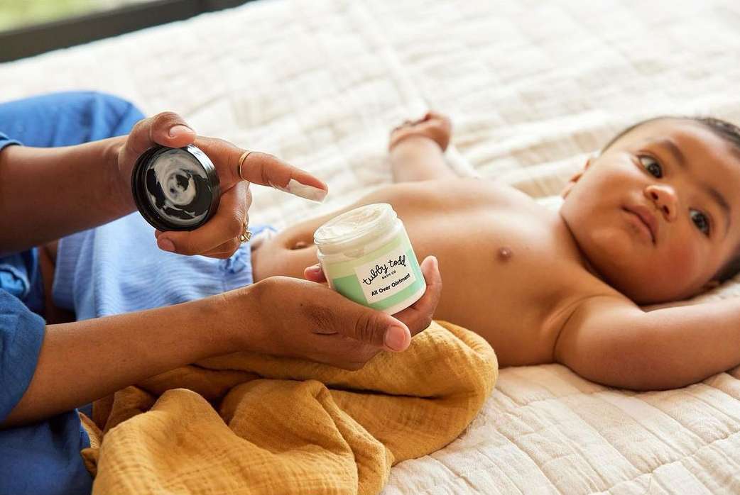 Close-up of a baby on a blanket with a person holding an open jar of Tubby Todd All Over Ointment and applying it to their finger.