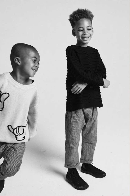 Two young children smiling in a black and white studio shot. One wears a white sweater; the other wears a black sweater and grey pants.