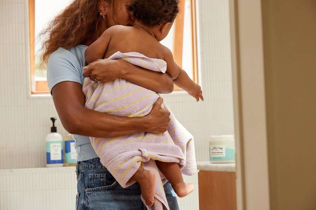 An adult woman holds a baby wrapped in a striped towel in a brightly lit bathroom with white tiled walls and toiletries.