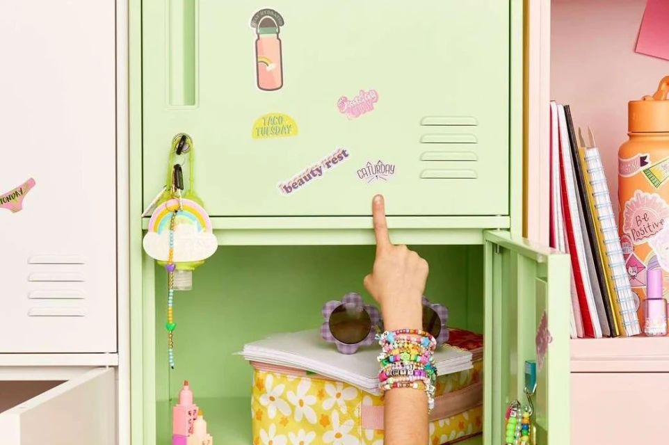 Child's hand points to a sticker on a mint green locker decorated with stickers, a rainbow keychain, and colorful beaded bracelets.