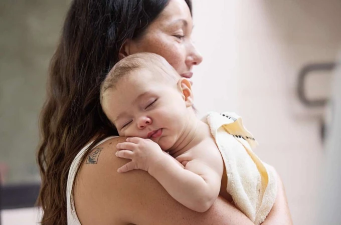A woman holds a sleeping baby against her shoulder, the infant wrapped in a soft towel in an indoor setting with natural lighting.