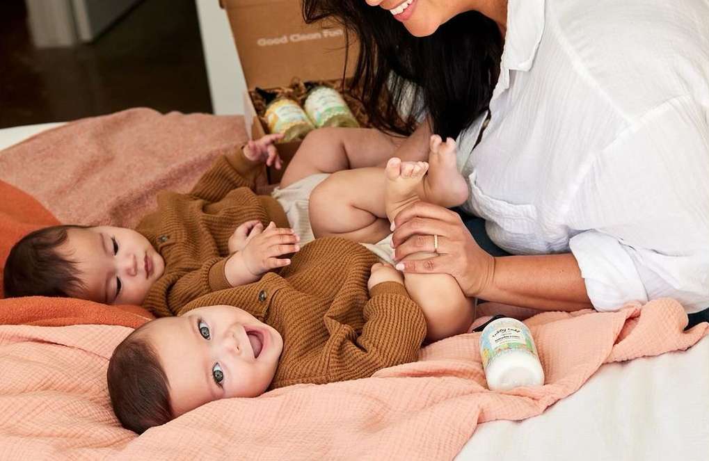 Two babies on a blanket with a parent, baby lotion, and product box in an indoor setting, showing elements of baby care and family life.