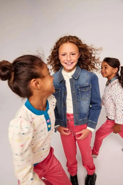 Three young girls in fashionable clothing smiling indoors. The girl in the center wears a denim jacket and pink pants, style shot, indoors.