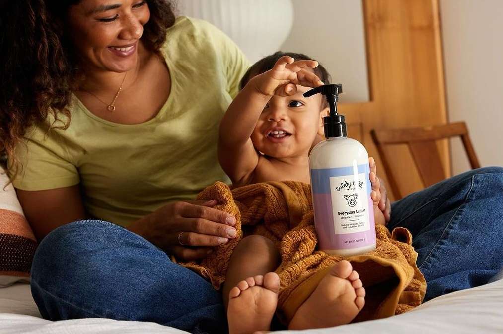 Smiling mother and baby on bed, holding a bottle of lotion; the infant is wrapped in a brown towel in an indoor setting.