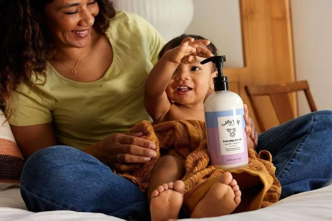 Smiling mother and baby on bed, holding a bottle of lotion; the infant is wrapped in a brown towel in an indoor setting.
