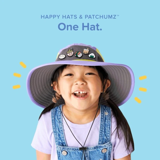 Smiling young child wearing a wide-brimmed hat with food-themed appliques in front of a soft blue background in a studio shot.