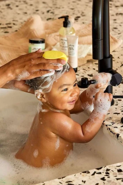 A baby smiles and is being washed with a yellow sponge by an adult in a bathroom with visible hair product bottles.