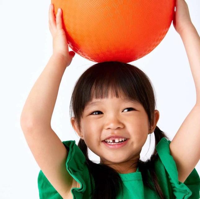 Adorable Asian child smiles, holding an orange ball on her head against a white background in a cheerful studio portrait.