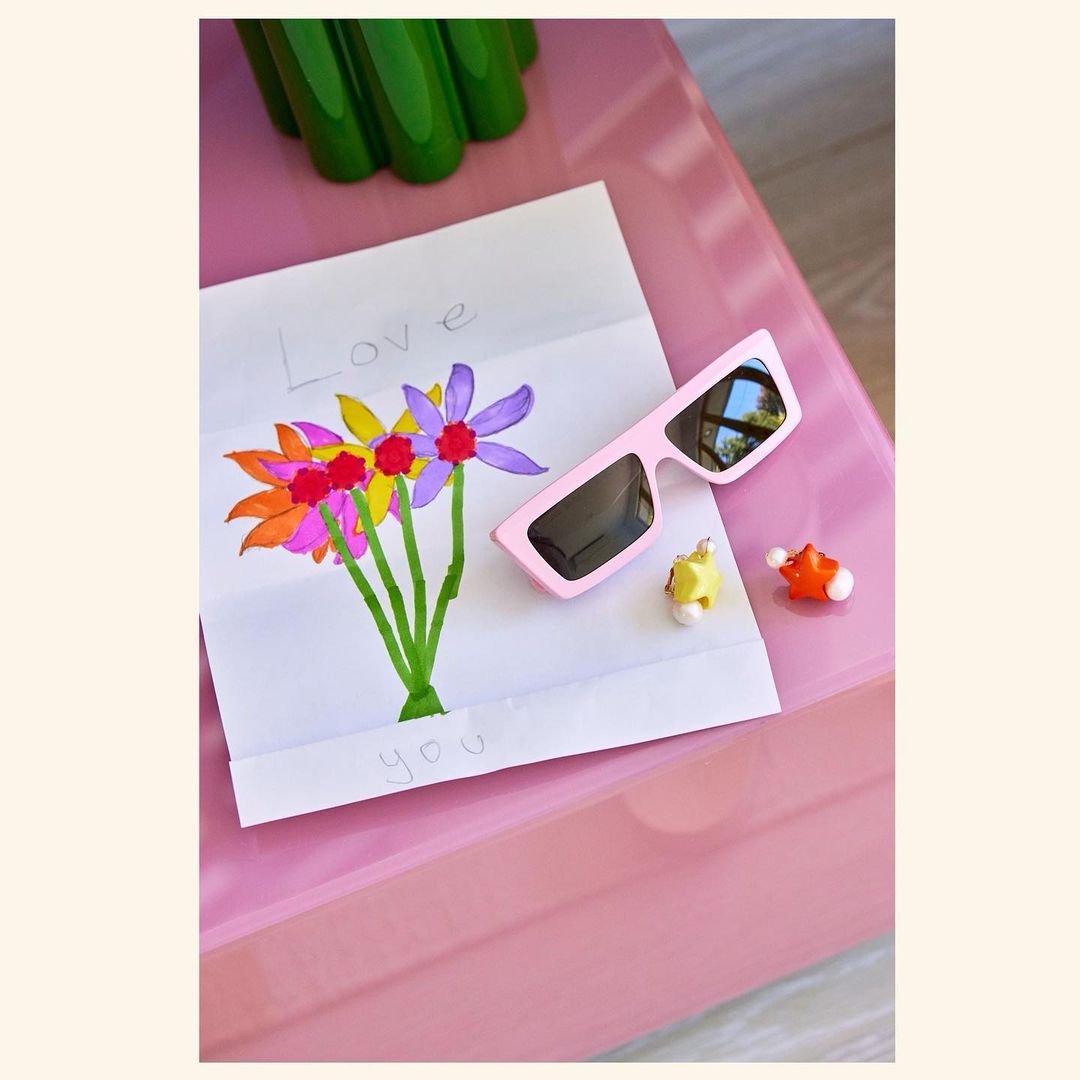 Still life composition on a pink surface featuring a child's flower drawing, pink sunglasses, and small animal figurines.