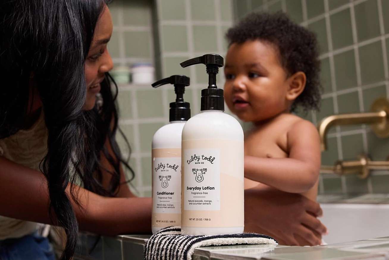 Mother and child near bathtub with two Tubby Todd bath products. Dark-haired woman watches her child near the bottles.