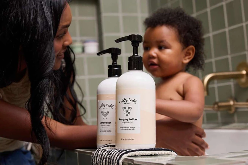 Mother and child near bathtub with two Tubby Todd bath products. Dark-haired woman watches her child near the bottles.