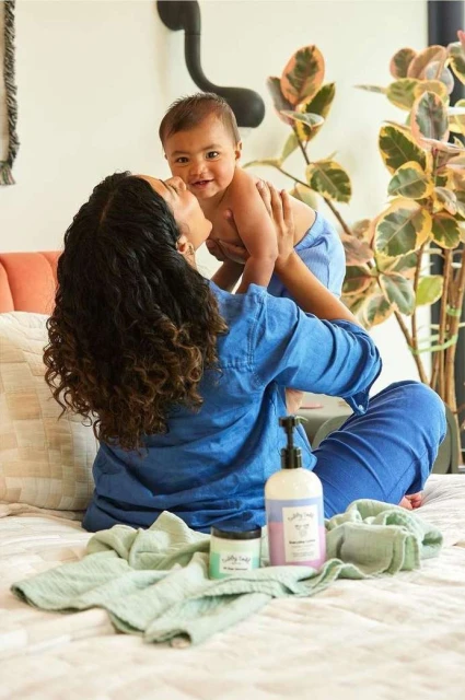 A mother in a blue shirt holds her baby in the air on a bed, with baby care products, indoors, under natural light near a houseplant.