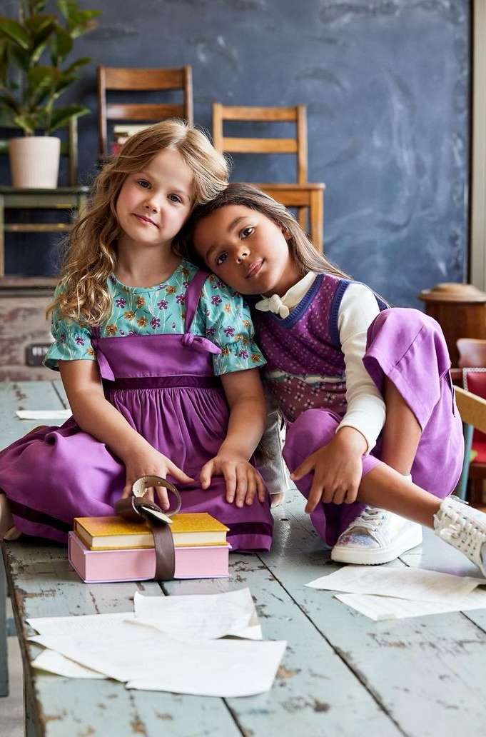 Two young girls in purple outfits sit at a table with books and papers. One girl rests her head on the other's shoulder.