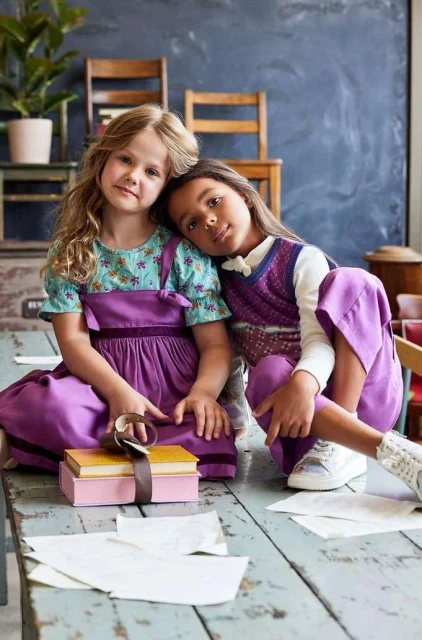 Two young girls in purple outfits sit at a table with books and papers. One girl rests her head on the other's shoulder.