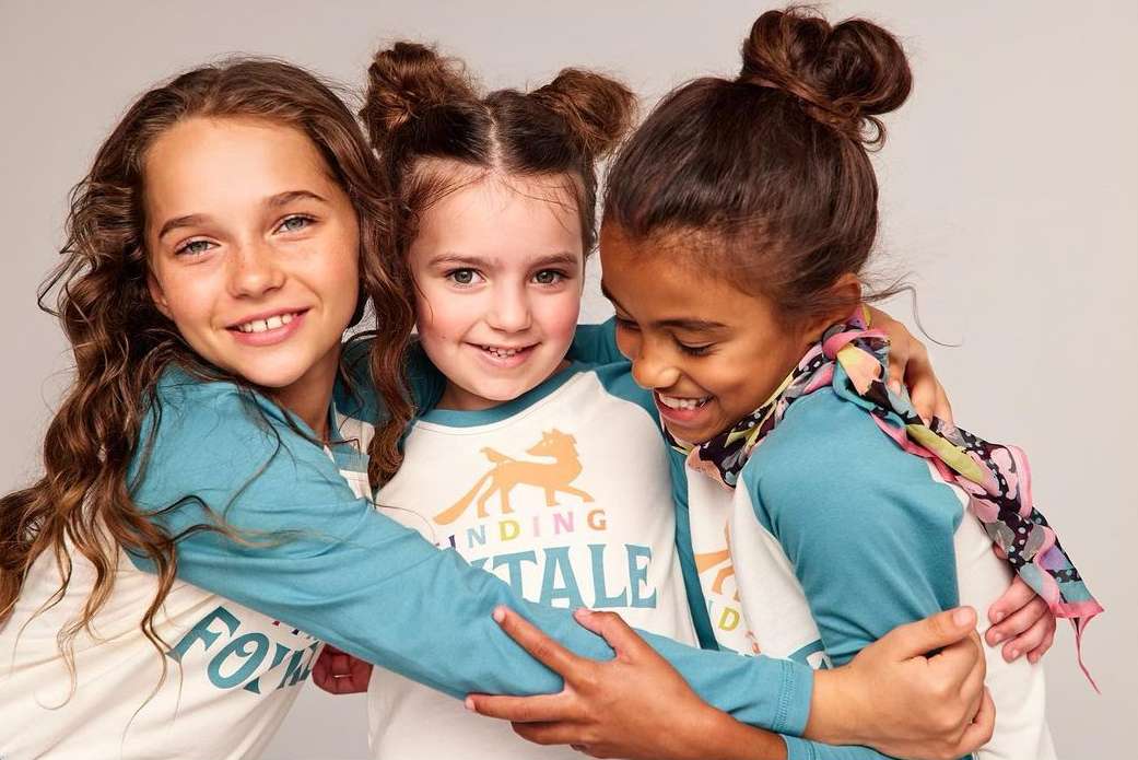 Three smiling young girls embrace closely in a studio setting, wearing matching shirts with a fox logo, capturing a joyful friendship.