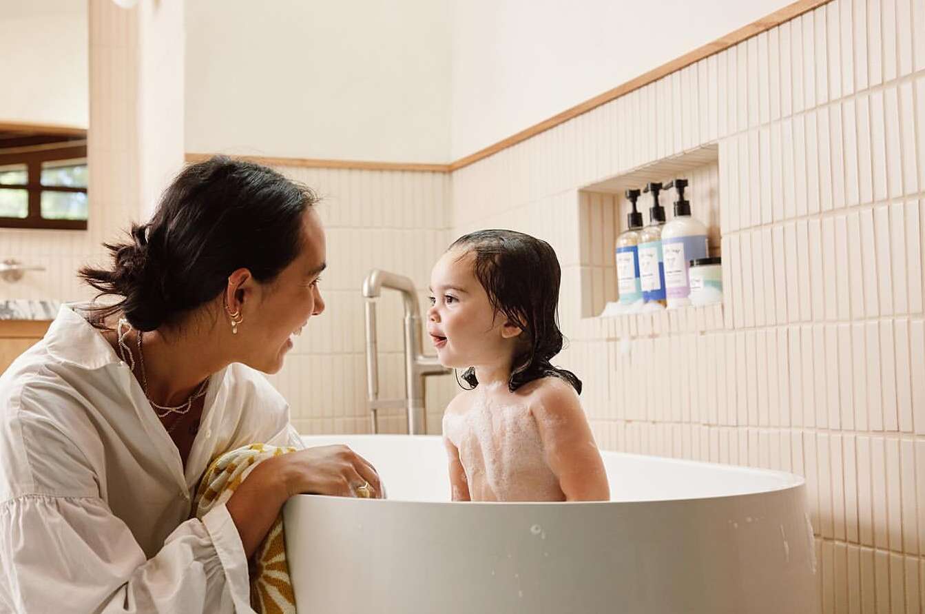 A child sits in a white bathtub filled with soapy water, a woman in a white shirt watches over her in an indoor bathroom setting.