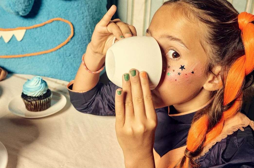 Young girl with star makeup and orange hair drinking from a white mug at a table with a frosted cupcake and a blue monster pillow.