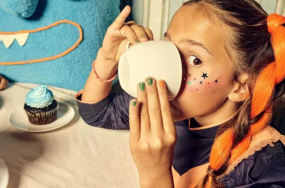 Young girl with star makeup and orange hair drinking from a white mug at a table with a frosted cupcake and a blue monster pillow.