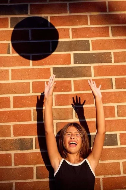 Girl stands in front of brick wall with arms raised, casting a circular shadow, laughing in bright sunlight, creating a cheerful portrait.