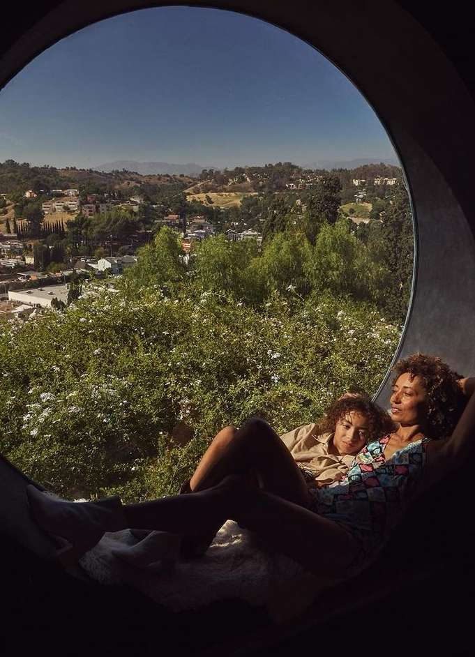 Two women lounge in a circular window overlooking a sunny landscape of trees and buildings in a residential area on a clear day.