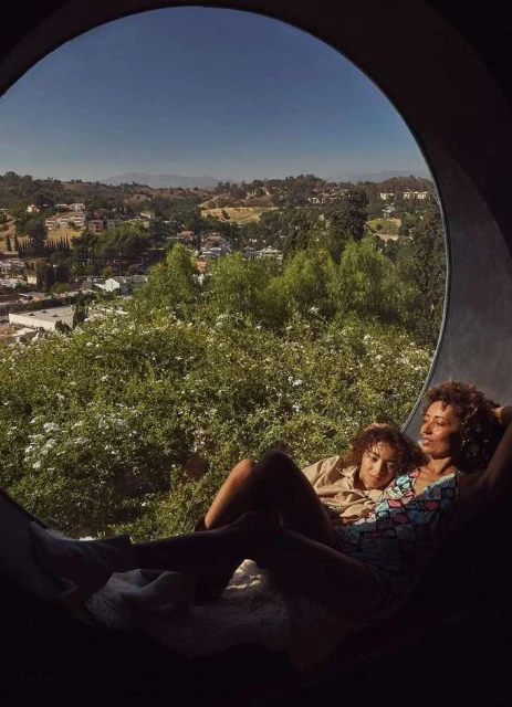 Two women lounge in a circular window overlooking a sunny landscape of trees and buildings in a residential area on a clear day.
