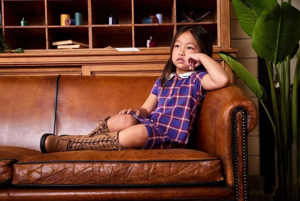 Young girl wearing a plaid dress and lace-up boots, sitting on a vintage leather couch in front of a wooden cabinet and potted plant.