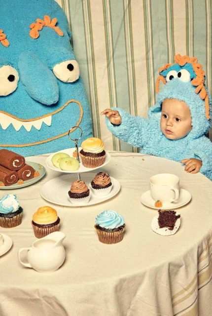 Child in a blue monster costume reaching for a cupcake at a table set with desserts and a white tablecloth; a blue monster pillow behind.