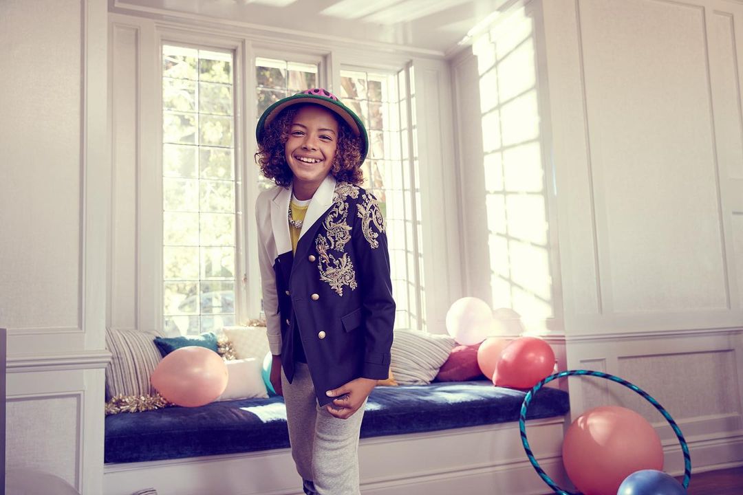 Portrait of a smiling child wearing a hat and an embellished blazer indoors near a window with balloons and a hula hoop.
