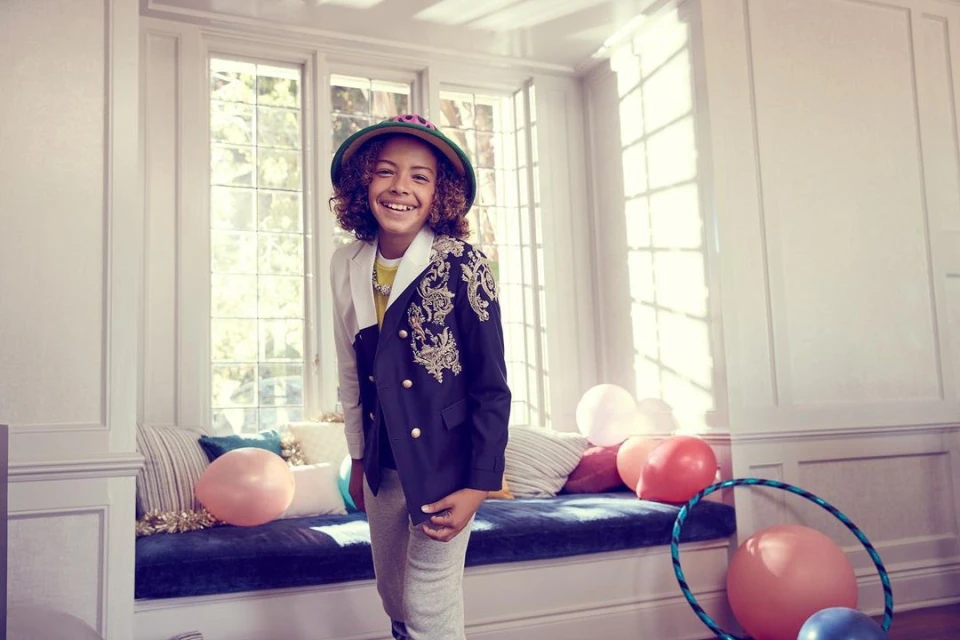 Portrait of a smiling child wearing a hat and an embellished blazer indoors near a window with balloons and a hula hoop.