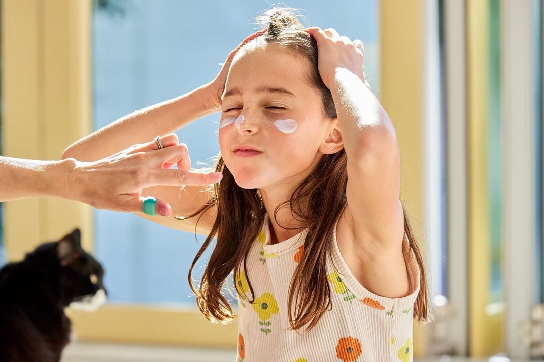Young girl with closed eyes smiles as cream is applied to her face by an adult hand in a brightly lit room with a black cat.