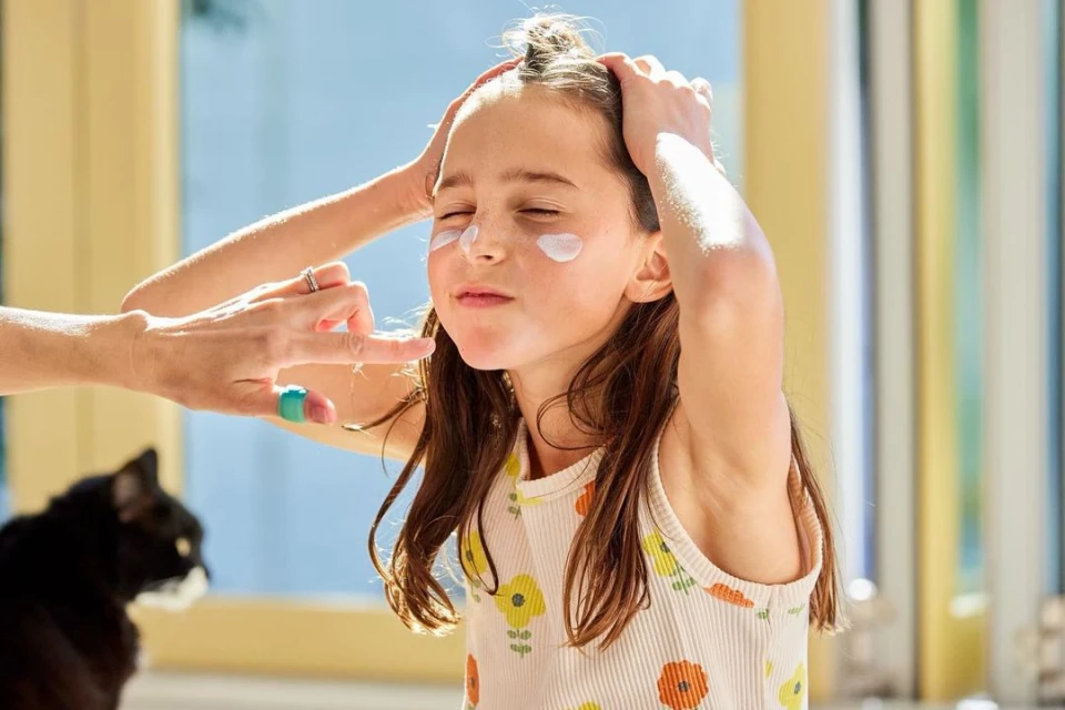 Young girl with closed eyes smiles as cream is applied to her face by an adult hand in a brightly lit room with a black cat.