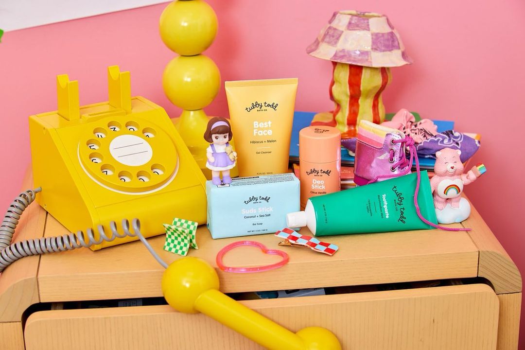 Cosmetic product arrangement on a wooden dresser with a yellow telephone, a lamp, and a Care Bear figurine against a pink wall.