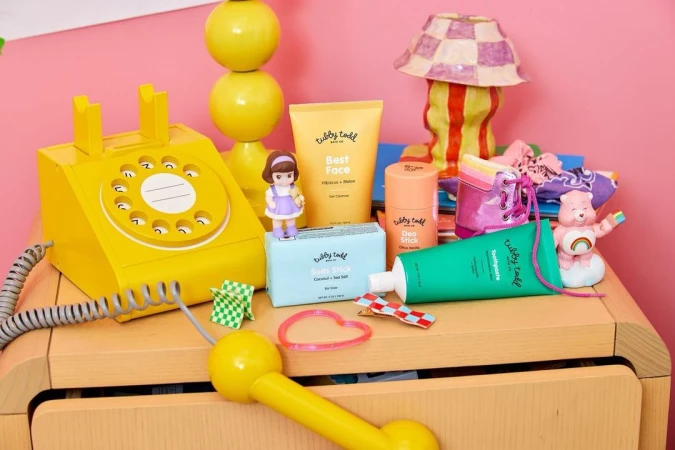 Cosmetic product arrangement on a wooden dresser with a yellow telephone, a lamp, and a Care Bear figurine against a pink wall.