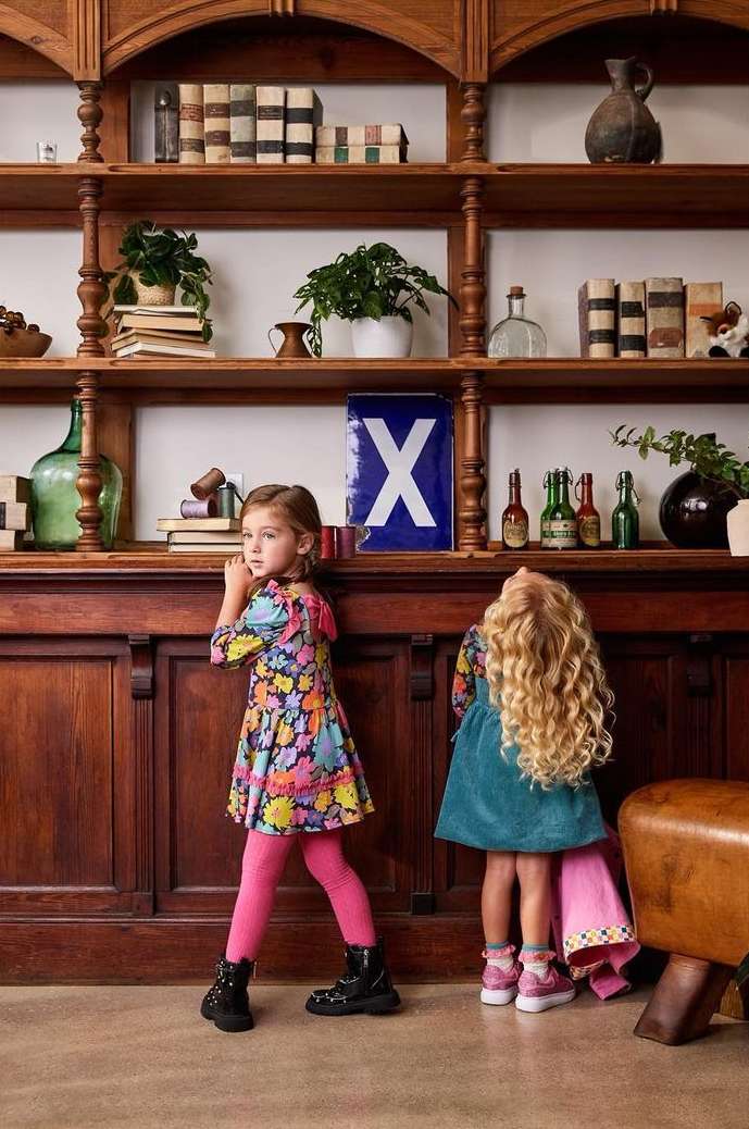 Two young girls stand in front of a dark wood shelf displaying books, plants, and vintage bottles inside of a home interior.