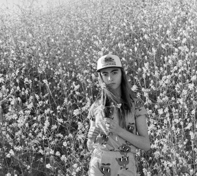 Young person wearing a California trucker hat and patterned t-shirt standing amidst a field of tall wildflowers.
